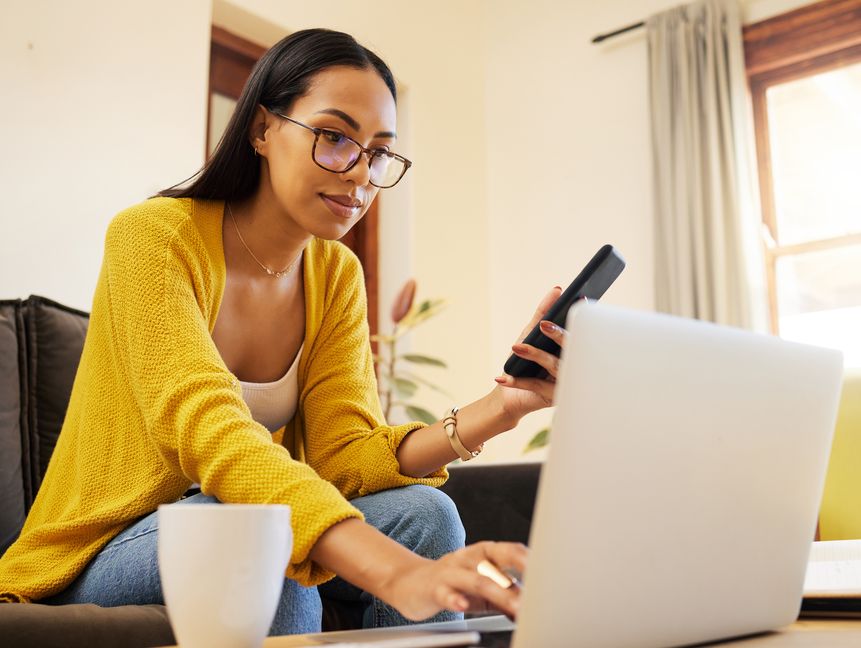 Woman sitting on a couch working on a laptop, holding a mobile phone 